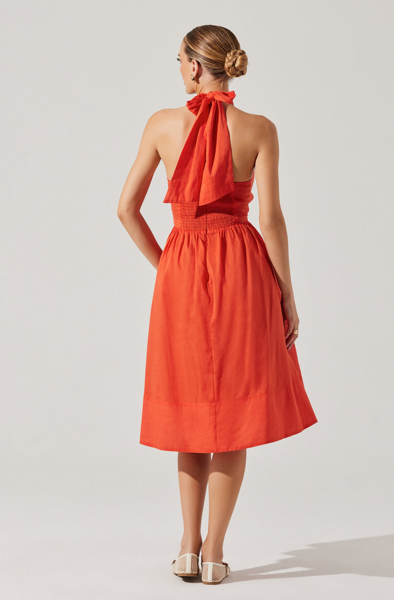 A woman is wearing a sleeveless, halter-neck dress in a bright orange color. The top part of the dress is fitted and gathered at the waist, and the skirt is full and knee-length with a slightly flared hem. The halter neckline features a large bow tied at the back of the neck, made from the same fabric as the dress. She is also wearing woven, open-toe shoes with a block heel.
