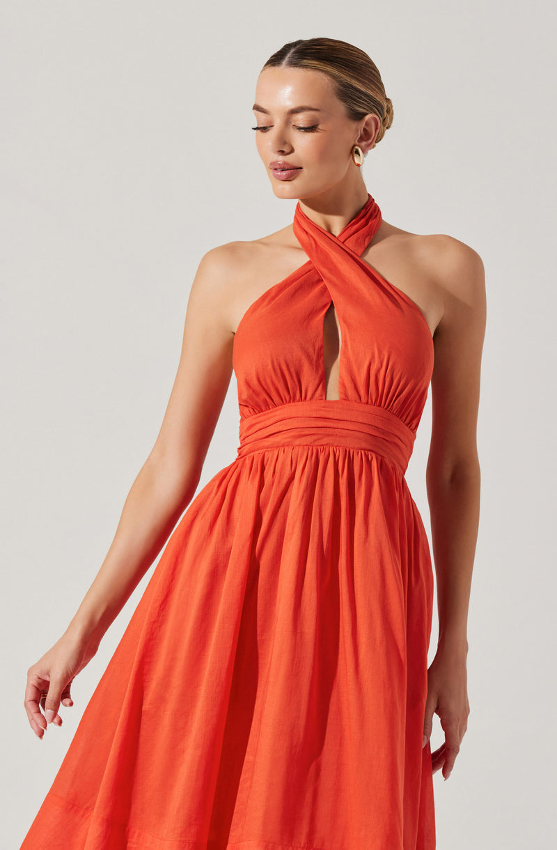 A woman is wearing a sleeveless, halter-style orange dress with a gathered waistband. The dress features a high neckline that twists around the neck, with a keyhole cutout in the front. The skirt is full and gathered, creating a flowing silhouette. The fabric appears to be lightweight and slightly textured.