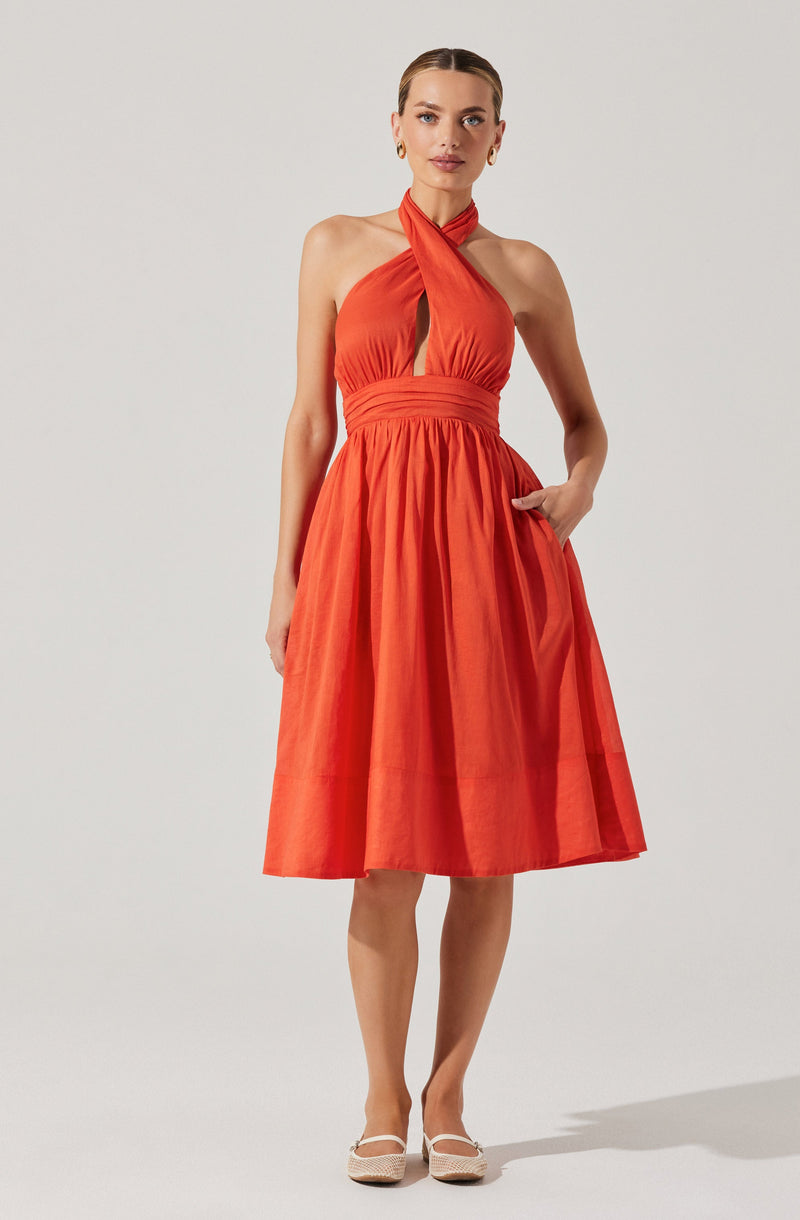 A woman is wearing a sleeveless, halter-neck dress in a bright orange color. The dress features a fitted bodice with a keyhole cutout at the front and an elastic waistband. The skirt is knee-length, full, and gathered, creating a voluminous silhouette. She has her right hand in a pocket on the side of the skirt and is wearing light-colored, closed-toe shoes with a strap.