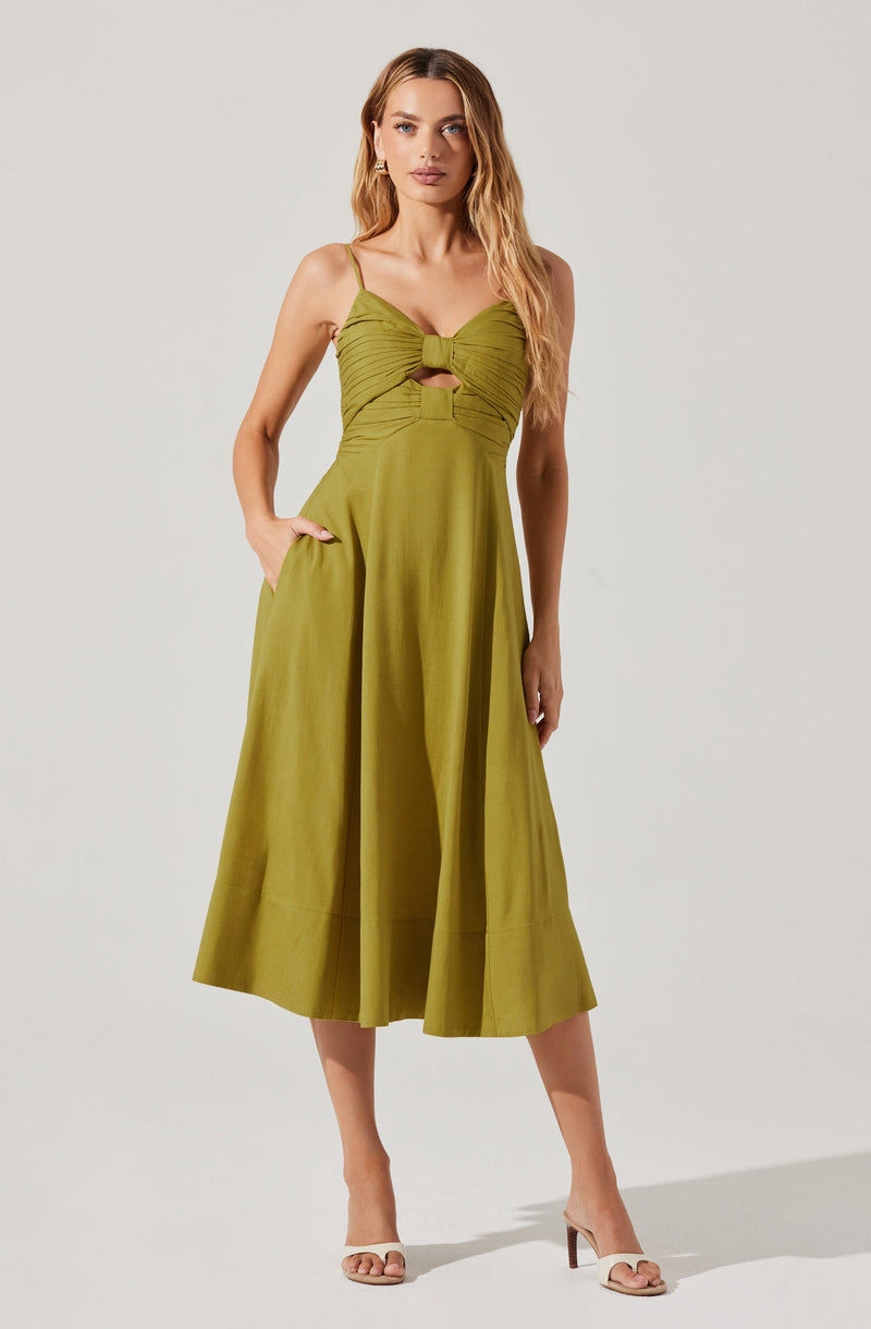 A woman is wearing a sleeveless, olive-green dress with spaghetti straps. The dress has a fitted bodice with a large, knotted detail at the center of the bust, creating a keyhole cutout below the knot. The skirt is full and flares out to mid-calf length, with a wide hem at the bottom. She completes the look with beige, open-toe high-heeled sandals.