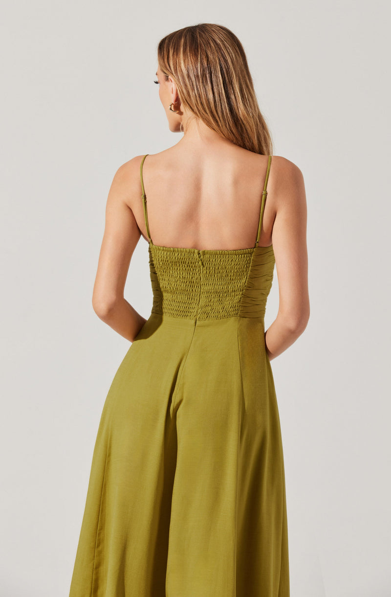A woman is wearing a sleeveless, olive green jumpsuit with thin spaghetti straps. The top part of the jumpsuit is a gathered, smocked fabric that fits snugly around her torso, while the bottom part consists of wide-leg pants made from a smooth, shiny material. The pants are high-waisted and flow down to the floor.