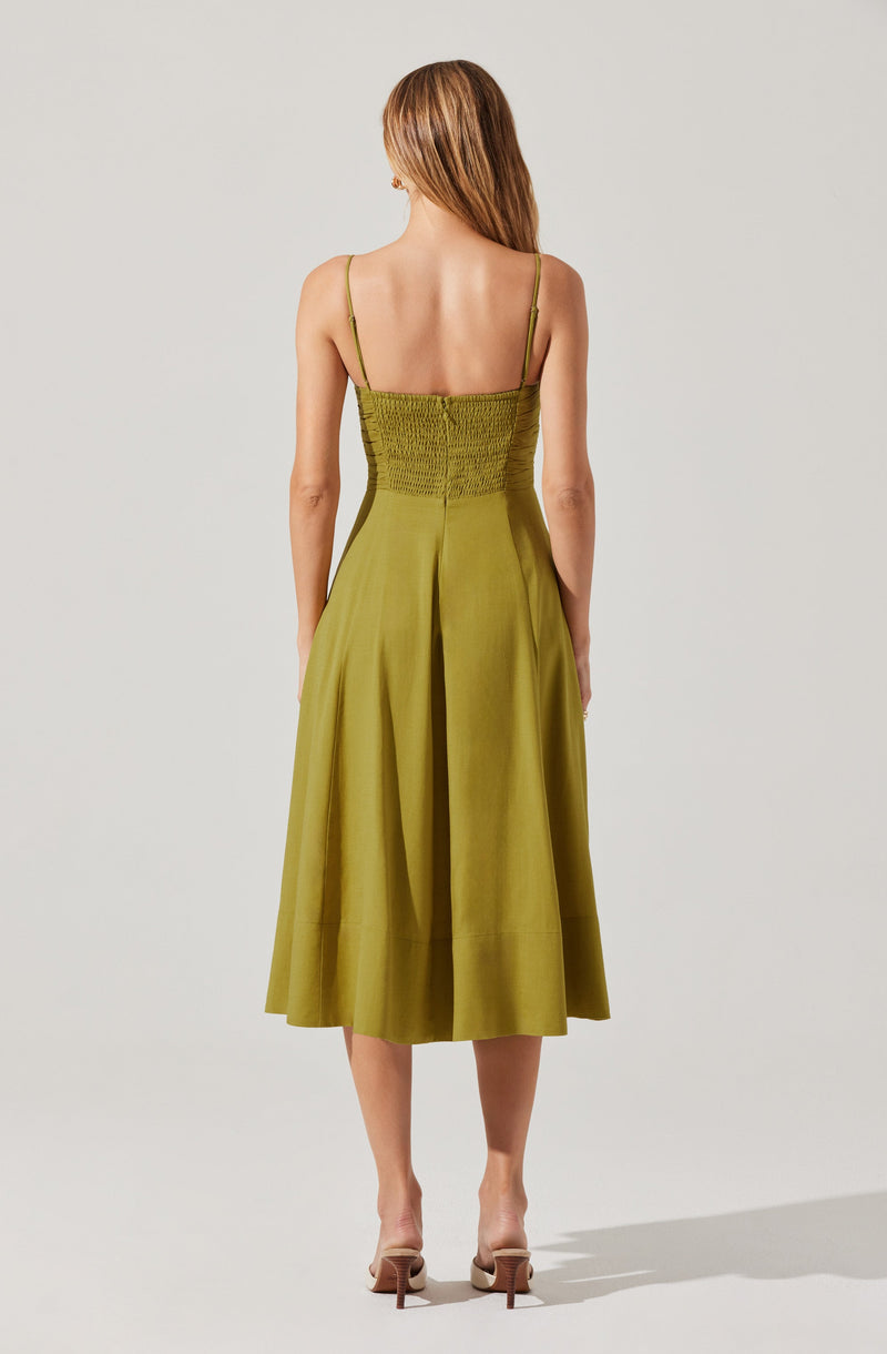 A woman is wearing a sleeveless, olive-green dress with thin spaghetti straps. The dress features a fitted, smocked bodice and a full, pleated skirt that falls to mid-calf length. She is also wearing nude-colored high-heeled sandals.