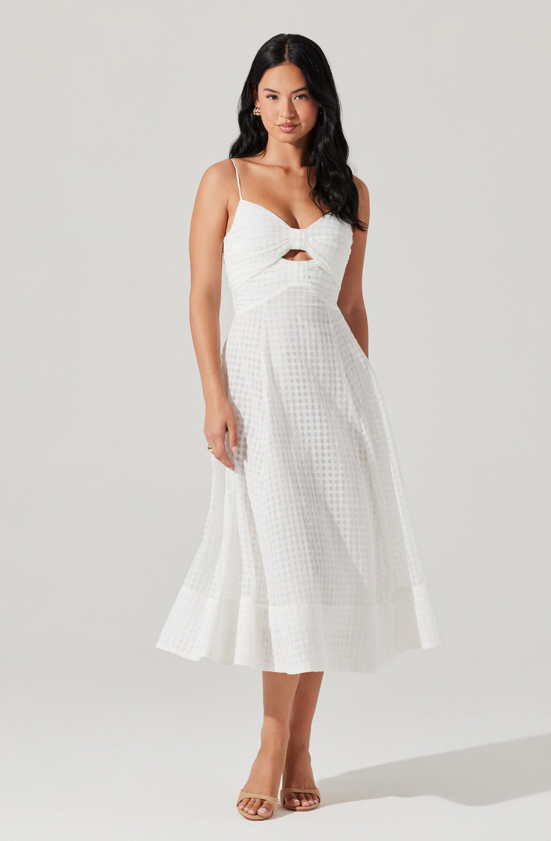 A woman is wearing a white, sleeveless dress with spaghetti straps. The dress features a fitted bodice with a ruched, bow-like detail at the chest and a small cutout below the bow. The skirt is high-waisted, flowing, and reaches mid-calf, with a subtle checkered pattern and a wide, pleated hemline.