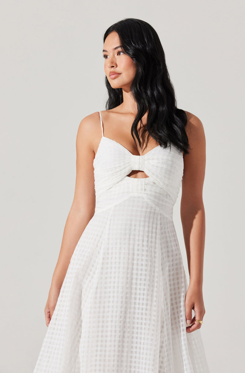 A woman is wearing a white sleeveless dress with thin spaghetti straps. The dress features a ruched bodice with a twisted knot detail at the center, creating a small cutout below the knot. The fabric has a subtle checkered pattern and flows down into a full skirt.