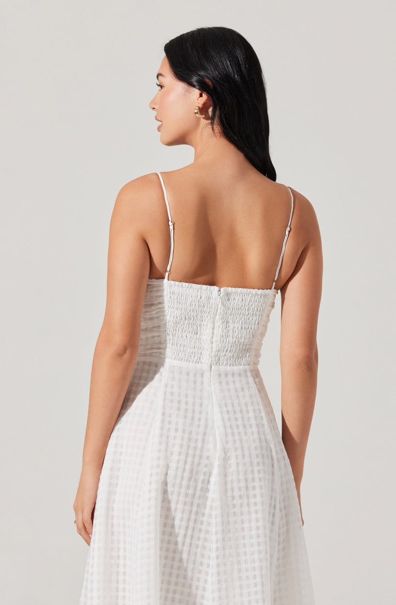 A woman is wearing a white, sleeveless dress with thin, adjustable spaghetti straps. The dress features a textured, smocked bodice and a high-waisted, flowing skirt with a subtle checkered pattern.