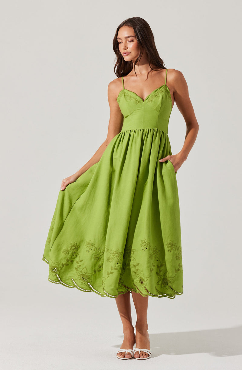 A woman is wearing a bright green dress with thin spaghetti straps. The dress features a fitted bodice with lace detailing and a full, flowy skirt with embroidered floral patterns near the hem. She is also wearing white high-heeled sandals.