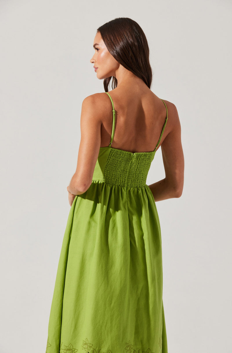 A woman is wearing a lime green sleeveless dress with thin adjustable straps. The bodice of the dress is fitted with a shirred or smocked section, and the skirt flows loosely with subtle pleats at the waist. The dress has a simple embroidered floral pattern near the hem.