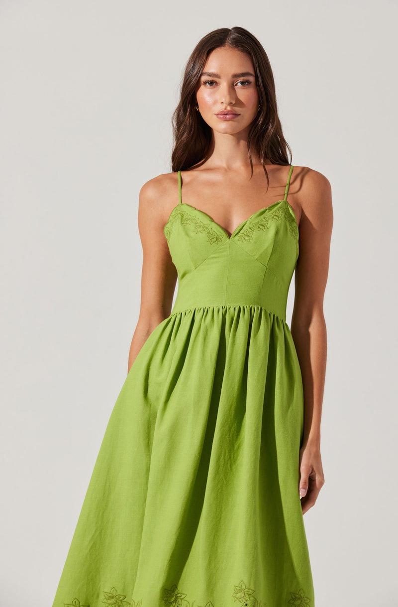 A woman is wearing a bright green dress with thin spaghetti straps. The dress features a fitted bodice with embroidered floral details along the neckline and a gathered, full skirt with matching embroidered floral patterns near the hem.