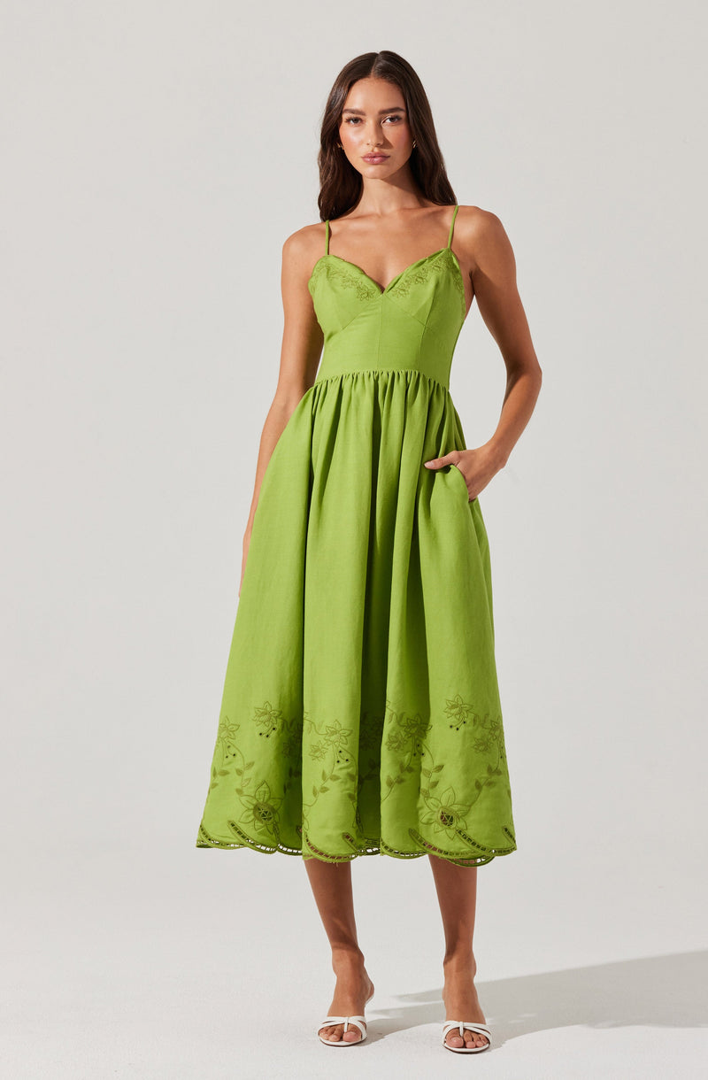 A woman is wearing a bright green sleeveless dress with spaghetti straps. The fitted bodice features embroidery details along the neckline. The skirt is gathered at the waist, flows to mid-calf length, and has floral embroidery along the hemline. The dress has pockets, and the woman is wearing white open-toe heels.