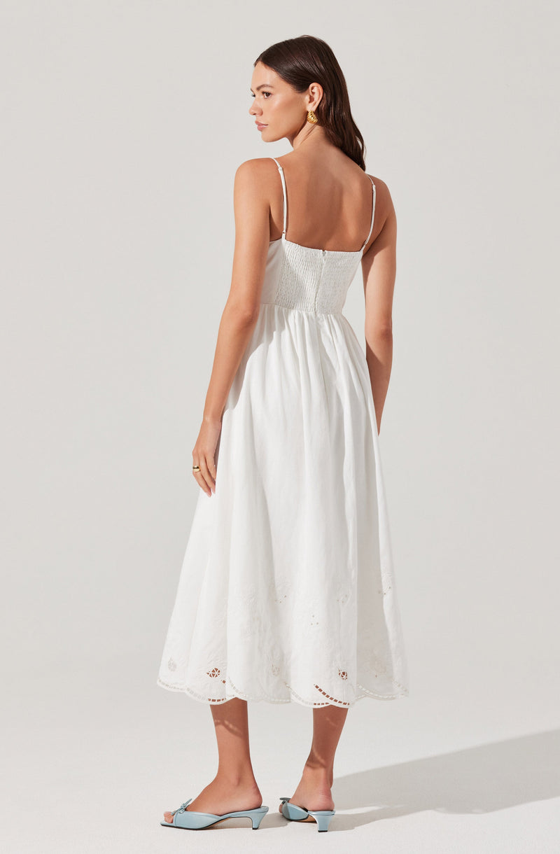 A woman is wearing a white, sleeveless, ankle-length dress with thin shoulder straps and embroidered detailing at the hem. She is also wearing matching light blue open-toe mules.