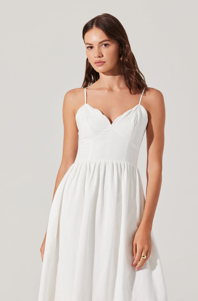 A woman is wearing a sleeveless white dress with thin spaghetti straps, a fitted bodice with subtle seam details, and a gathered, voluminous skirt that falls to the floor.