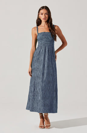 A woman is wearing a blue sleeveless maxi dress with adjustable straps. The dress features a smocked bodice with a fitted waist and a long, flowing skirt made of textured, crinkled fabric. She is also wearing tan high-heeled sandals.