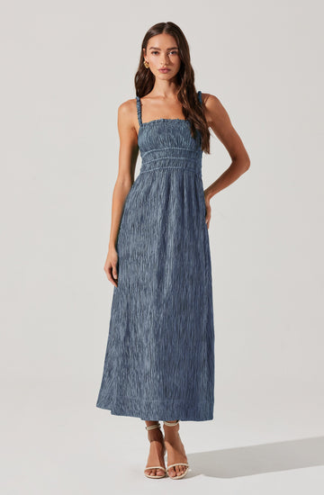 A woman is wearing a blue sleeveless maxi dress with adjustable straps. The dress features a smocked bodice with a fitted waist and a long, flowing skirt made of textured, crinkled fabric. She is also wearing tan high-heeled sandals.