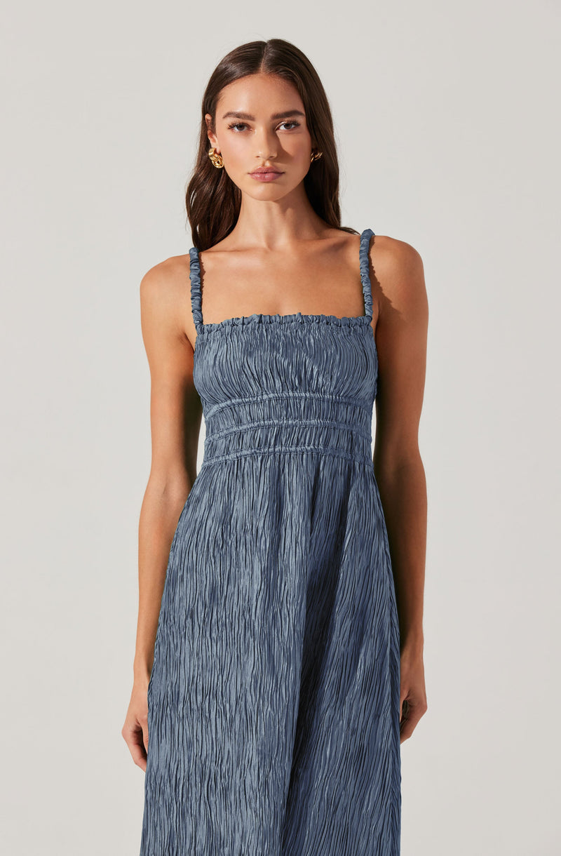A woman is wearing a sleeveless, blue dress with ruched fabric throughout. The dress features thin, ruffled shoulder straps and an elasticized, fitted bodice. The fabric has a textured, crinkled appearance, creating an elegant and relaxed look.
