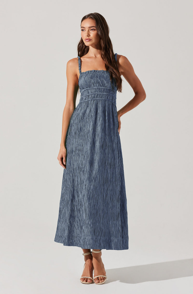 A woman is wearing a sleeveless, full-length, blue crinkled maxi dress with thin straps. The dress has a fitted bodice with shirred detailing and a flowing skirt. She is also wearing strappy beige high-heeled sandals.