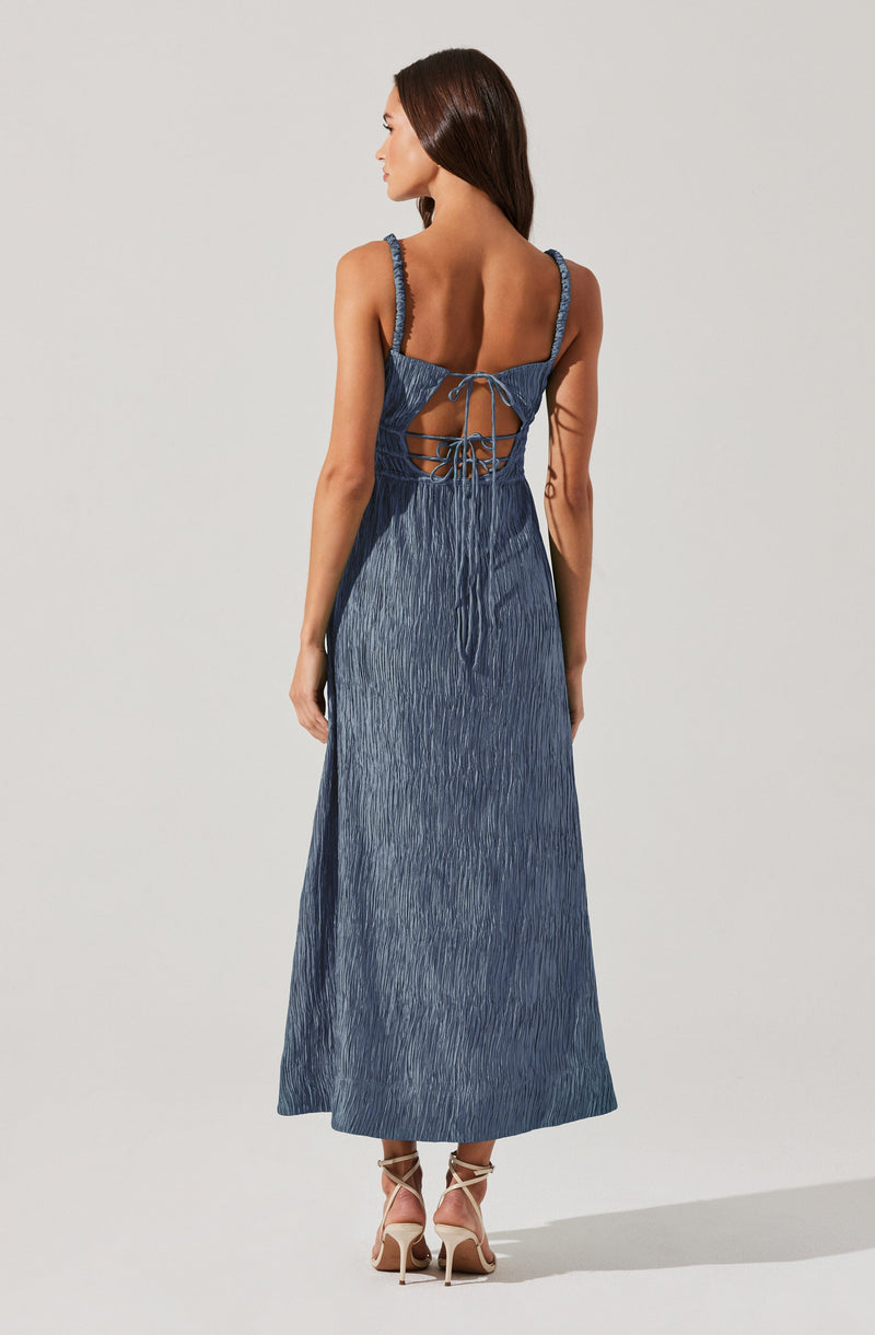 A woman is wearing a long, sleeveless denim dress with textured fabric. The dress features braided shoulder straps and an open back with crisscross laces tied in a bow. The hem reaches down to her ankles, and she is wearing high-heeled sandals with ankle straps.