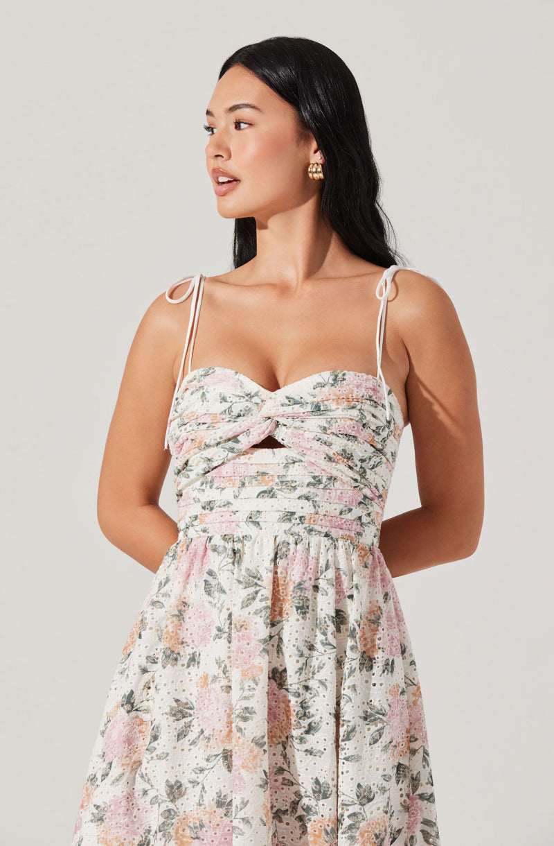 A woman is wearing a sleeveless dress with thin shoulder straps tied into bows. The dress features a fitted bodice with a sweetheart neckline and a center knot detail. The skirt part of the dress is flowy and has a floral pattern with soft pastel colors, including pink, green, and peach, on a light background.