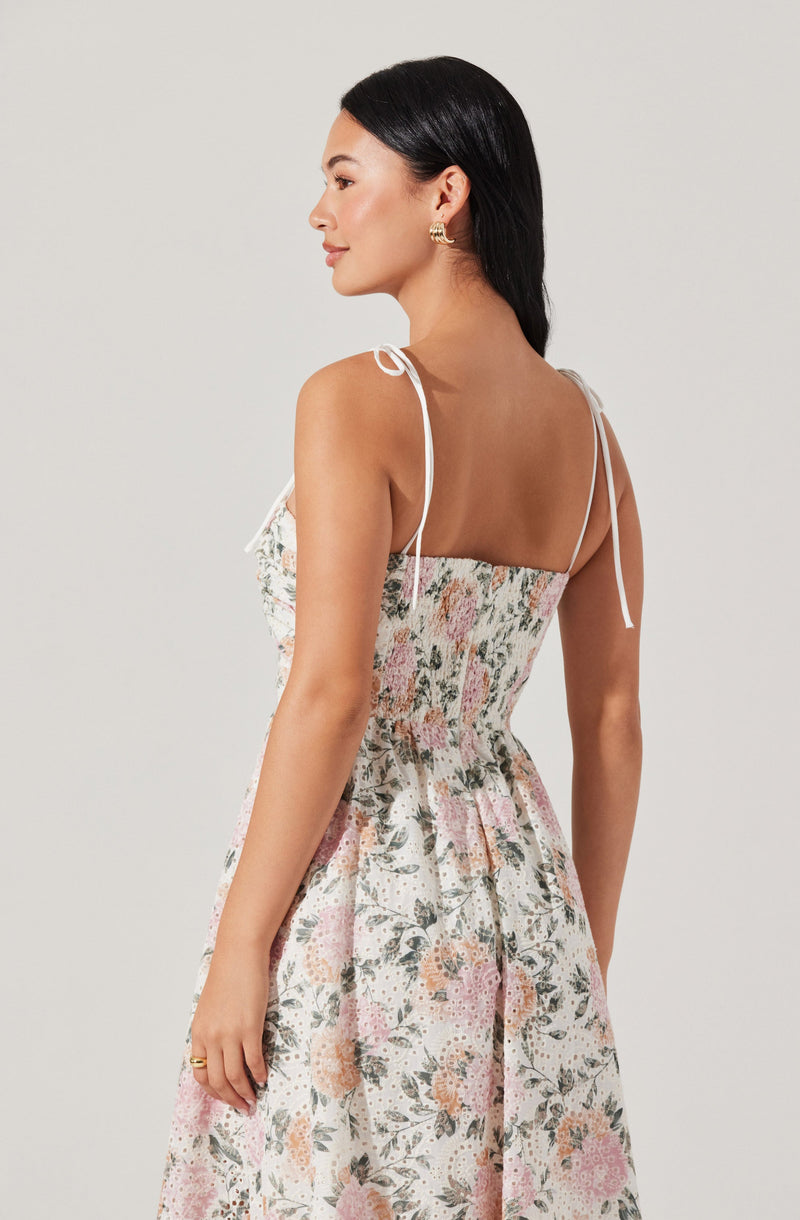 A woman is wearing a sleeveless, floral-print dress with thin, white spaghetti straps tied into bows on her shoulders. The dress features a fitted bodice with a smocked, elastic waistband and a flowing skirt with a pink and green floral pattern on a light background. She has short, dark hair and is accessorized with gold hoop earrings and a gold ring.