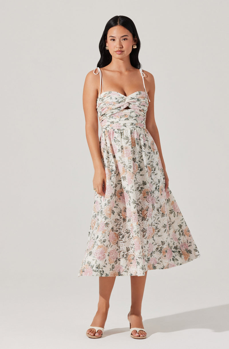 A woman is wearing a sleeveless, midi-length dress with thin spaghetti straps tied at the shoulders. The dress features a fitted bust with a twist knot detail at the center and a gathered waist, flowing into a full, floral-patterned skirt. The fabric has a light, soft color palette with pink, green, and white shades and a botanical print. She is also wearing open-toed, heeled sandals and gold earrings.
