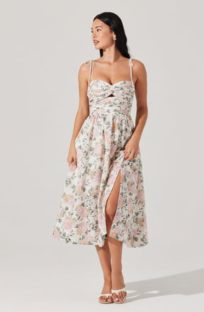 A woman is wearing a sleeveless dress with thin off-shoulder straps tied into bows on her shoulders. The dress has a fitted bodice with a twisted knot detail at the bust and a cutout below it. The skirt is long, flowy, and features a floral print with soft pastel colors. There is a slit on the front of the skirt revealing part of her leg. She is wearing open-toe sandals and has her dark hair down.