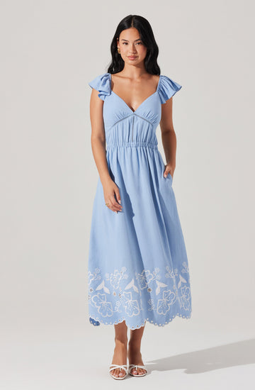 A woman is wearing a light blue, sleeveless, V-neck maxi dress with ruffled shoulder details. The dress has a gathered waistline and features an embroidered floral and scalloped hem pattern in white at the bottom. She is also wearing white open-toed heels.