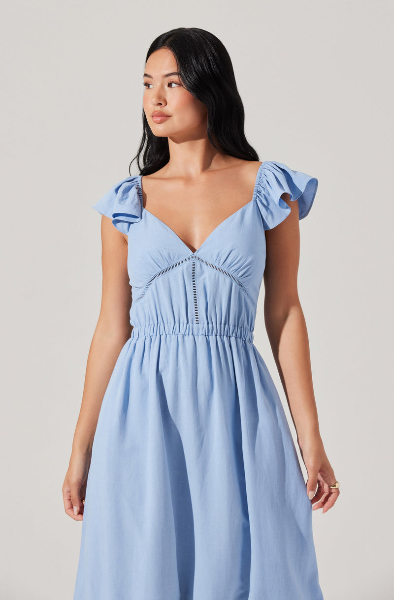 A woman is wearing a light blue dress with a V-neckline and short, ruffled sleeves. The dress features a smocked waistband and stitched detailing along the bust area, creating a fitted silhouette at the top and a flowy skirt.