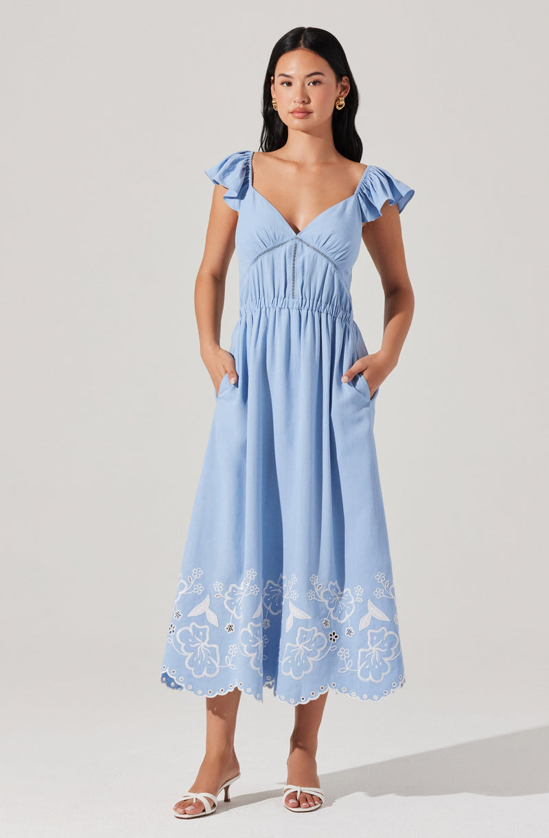 A woman is wearing a light blue, midi-length dress with ruffled cap sleeves, a V-neckline, and a fitted waist. The skirt of the dress features floral embroidery near the hemline. She has paired the dress with white strappy heeled sandals and gold hoop earrings.