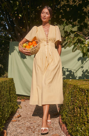 A woman is wearing a light yellow, ankle-length dress with short, puffed sleeves and a deep V neckline. The dress features a gathered, ruched bust with decorative stitching and a tie detail at the waist. She is holding a wicker basket filled with assorted fruits, including oranges, in her left hand. The background consists of green foliage and a light green cloth hanging, with sunlight casting shadows on her and the surroundings.