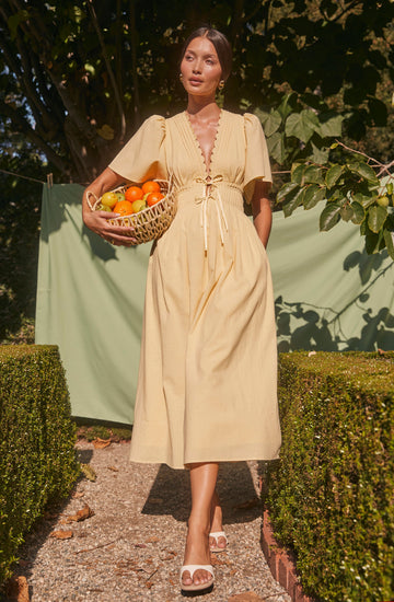 A woman is wearing a light yellow, ankle-length dress with short, puffed sleeves and a deep V neckline. The dress features a gathered, ruched bust with decorative stitching and a tie detail at the waist. She is holding a wicker basket filled with assorted fruits, including oranges, in her left hand. The background consists of green foliage and a light green cloth hanging, with sunlight casting shadows on her and the surroundings.