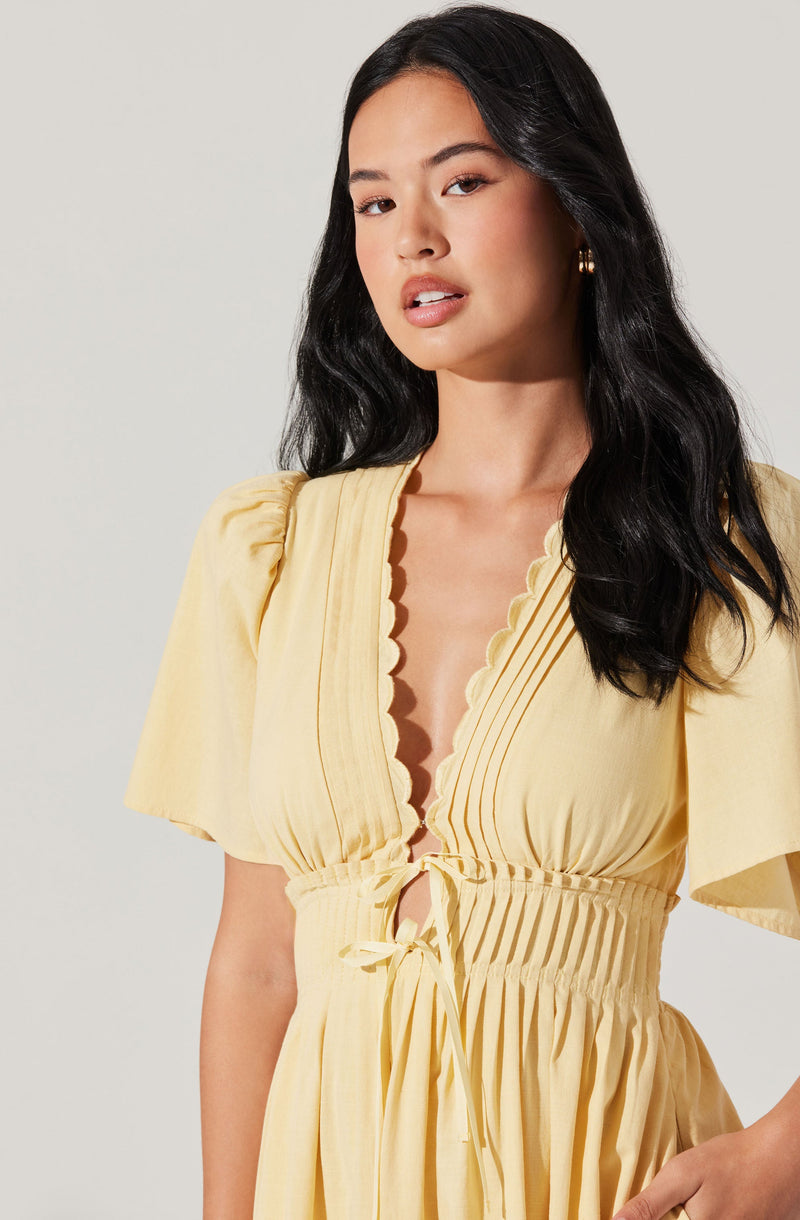 A woman is wearing a light yellow dress with short puffed sleeves. The dress features a deep V neckline with a scalloped trim, and there are vertical pleats along the bodice. The waistline is cinched with a gathered elastic band and a matching fabric tie at the front.