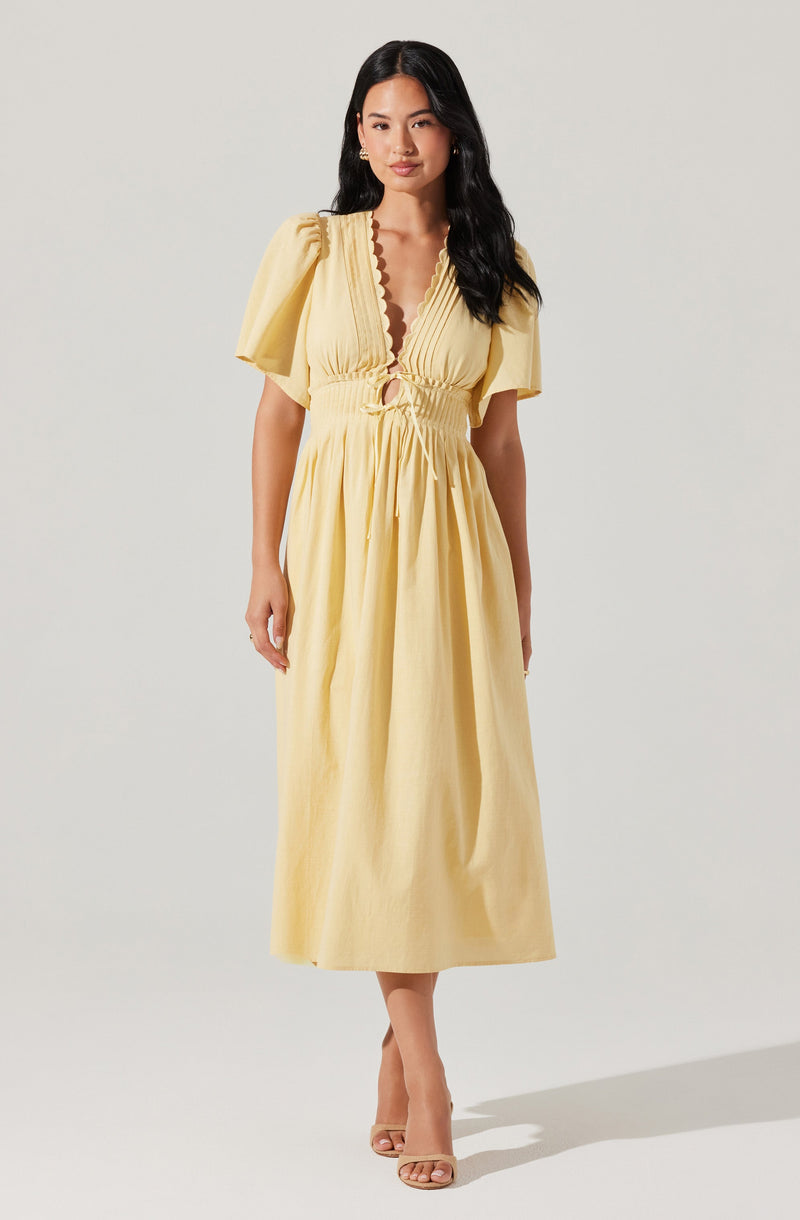 A woman is wearing a yellow maxi dress with puffed short sleeves, a deep V neckline with a scalloped edge, and a gathered, ruched waist with a tie-up detail. The dress has a flowing skirt that reaches the ankles.