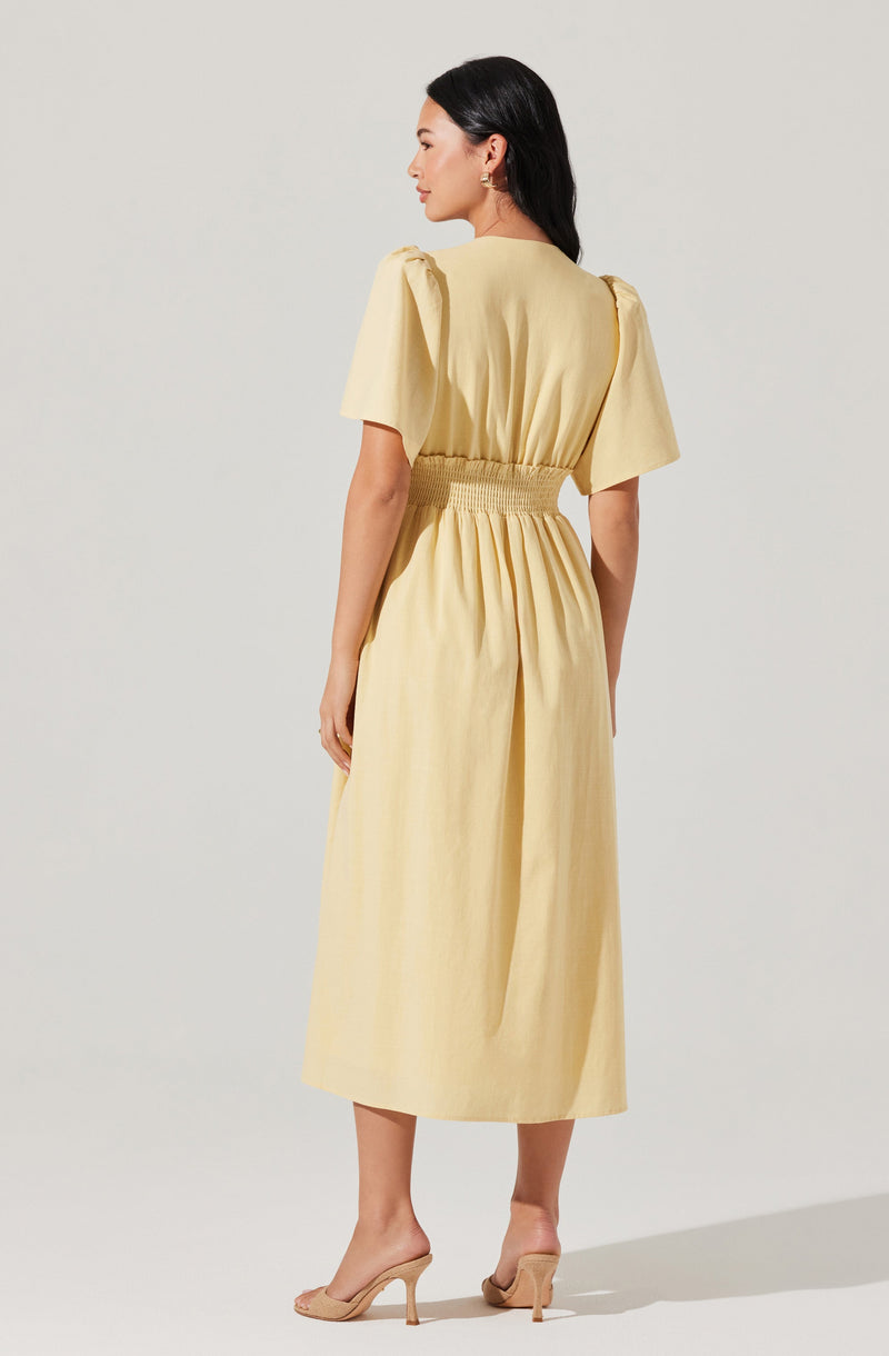 A woman is wearing a light yellow dress with puffed short sleeves. The dress has a fitted, elasticized waist and a long, flowing skirt that reaches mid-calf. She is also wearing nude high-heeled shoes.