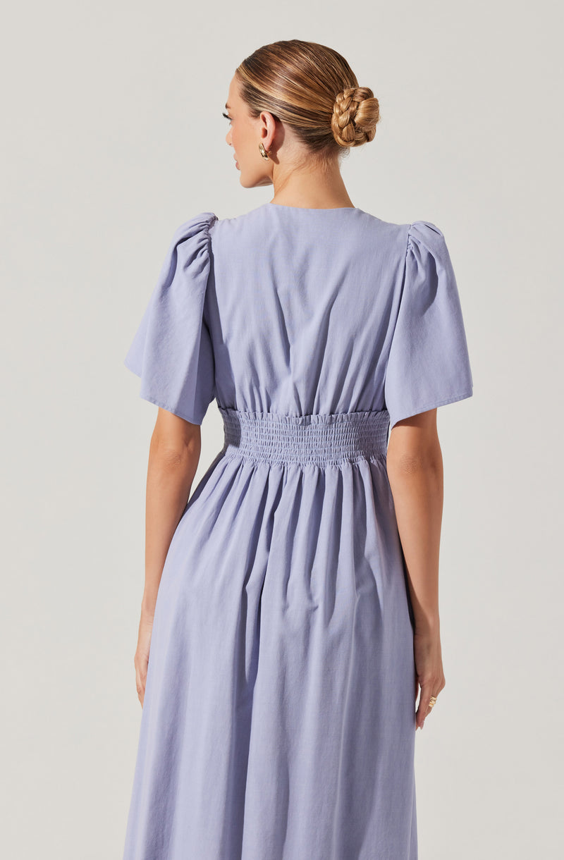 A woman is wearing a light lavender-colored dress with puffed short sleeves and a fitted, elasticized waistband. The fabric appears lightweight and has a smooth texture. The dress flows loosely from the waist down. She has her hair styled in a low bun and is wearing earrings.