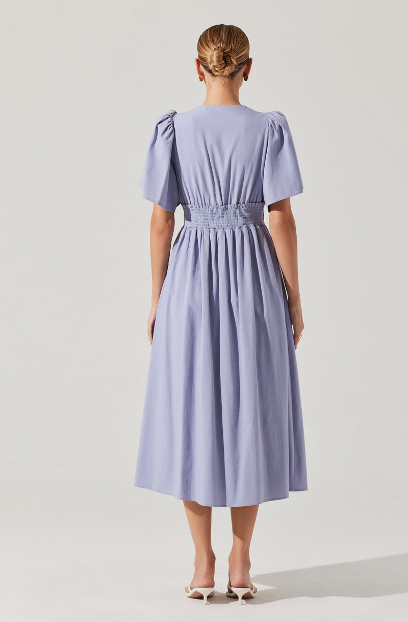 A woman is wearing a light blue, midi-length dress with short puffed sleeves. The dress features a high, gathered waistband that creates a slightly cinched waist, and the skirt has soft gathers, giving it volume and a flowing appearance. She is also wearing white high-heeled shoes.