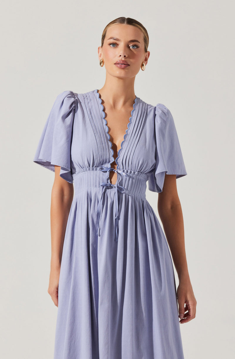 A woman is wearing a light purple dress with puffed short sleeves, a deep V-neckline edged with scalloped trim, and a gathered waist with a tied bow. The dress has a flowing, floor-length skirt.