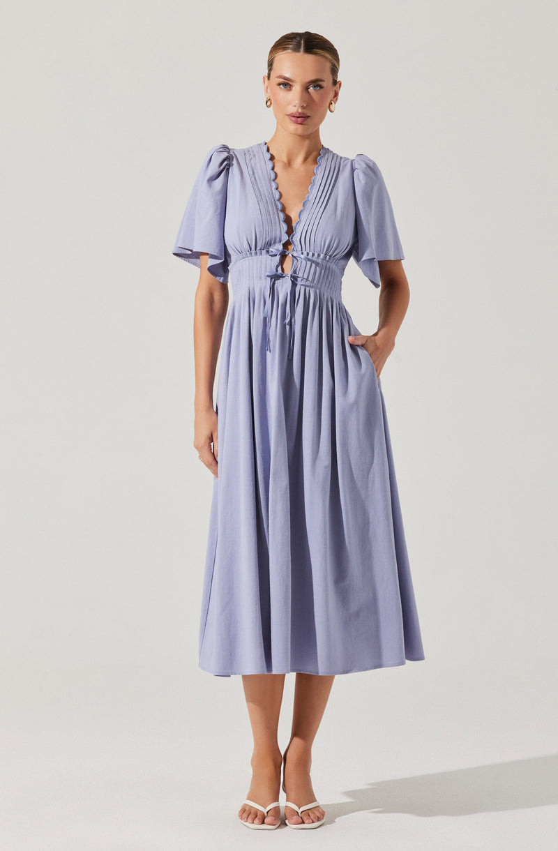 A woman is wearing a light purple, midi-length dress with a deep V-neckline edged with scalloped trim. The dress has puffed, short sleeves and a fitted waist with a drawstring detail at the front. The skirt is gathered and flows down to her mid-calf. She completes her look with white high-heeled sandals and gold hoop earrings.