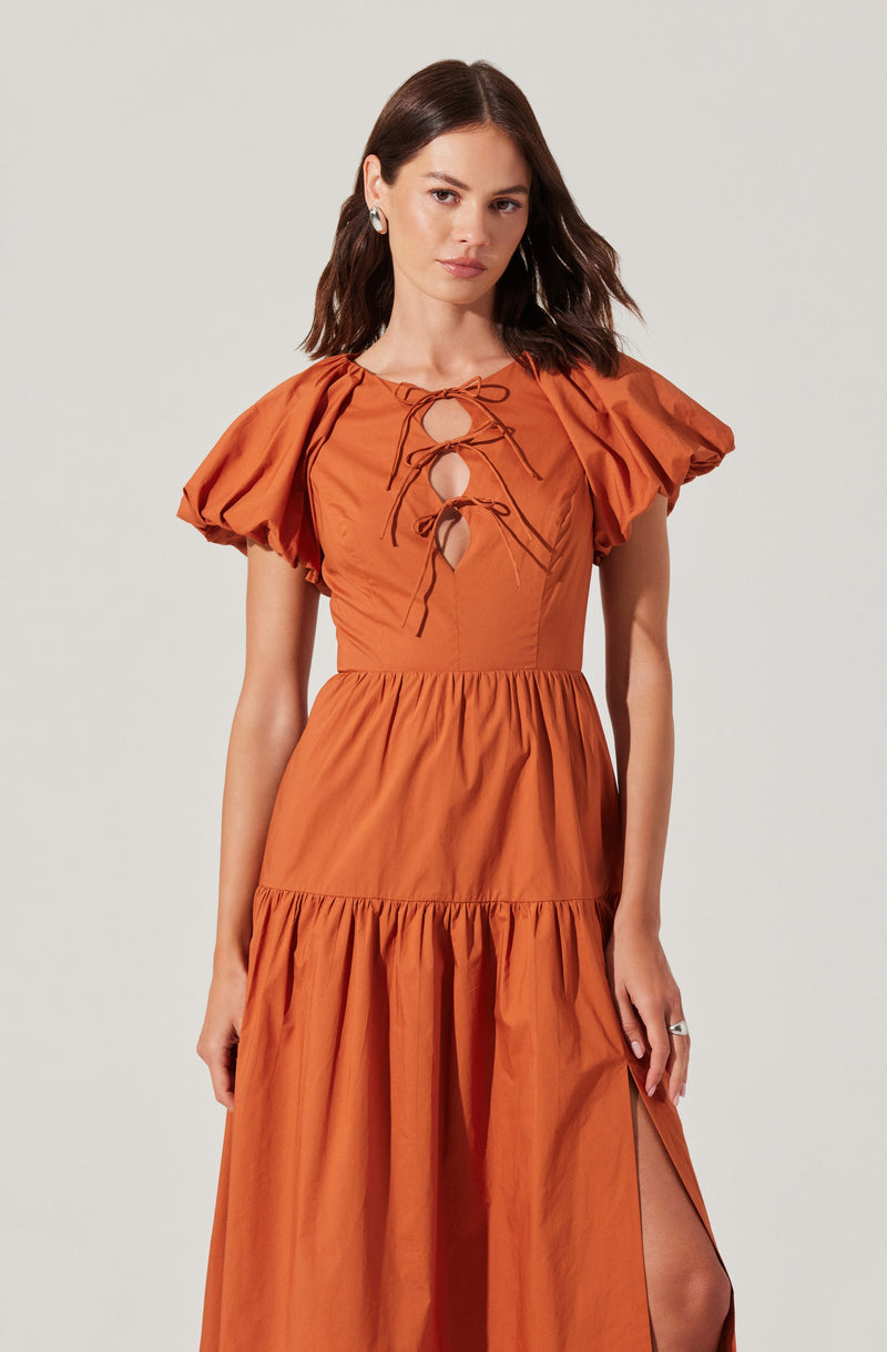 A woman is wearing a coral-colored dress with puffed short sleeves, a fitted bodice with a lace-up front featuring multiple bows, and a tiered skirt with a slit on one side.