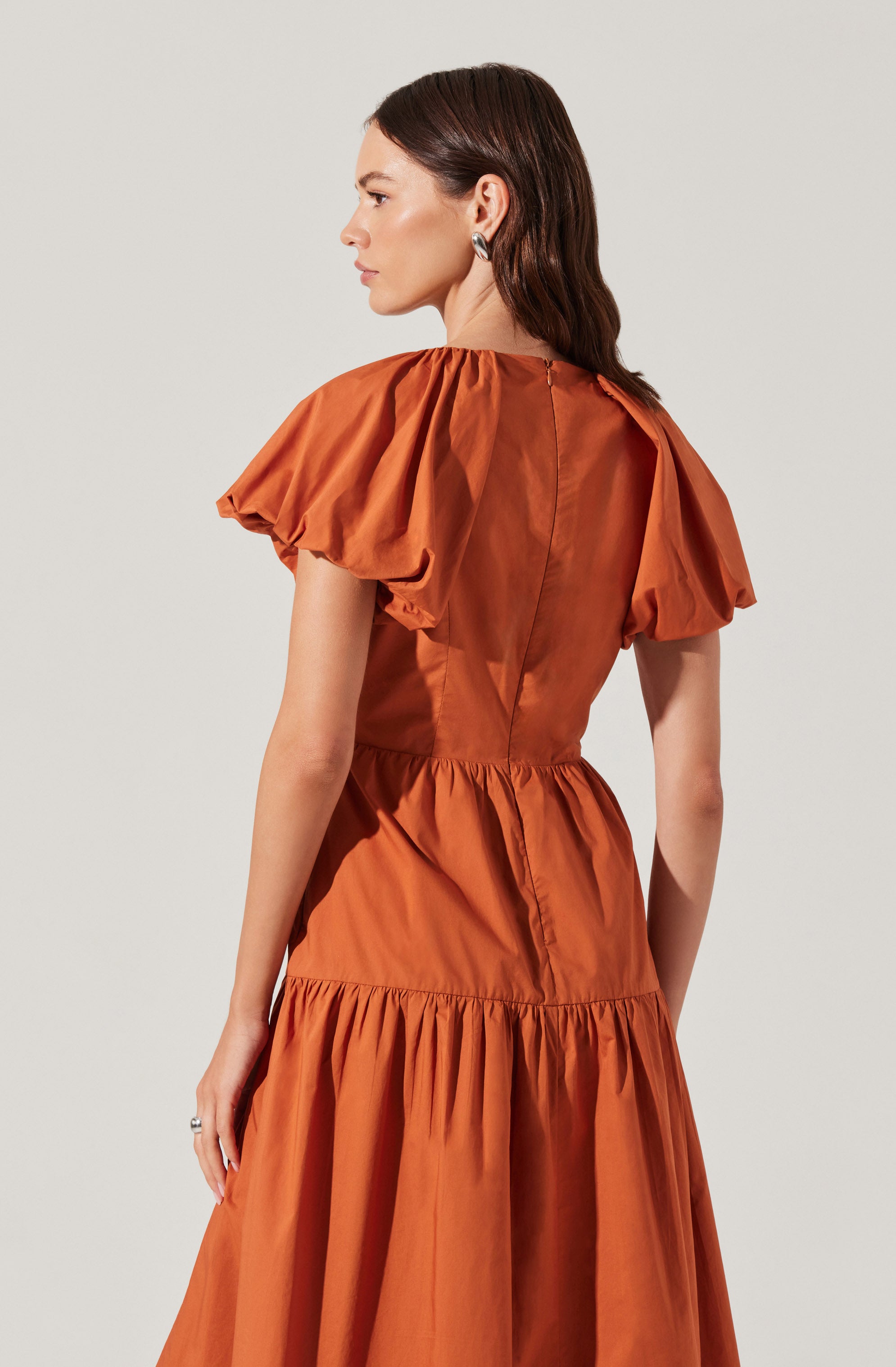 A woman is wearing a rust-colored dress with short, puffed sleeves. The dress has a fitted waist and a tiered skirt with gathered fabric. The material appears to be lightweight and slightly shiny.