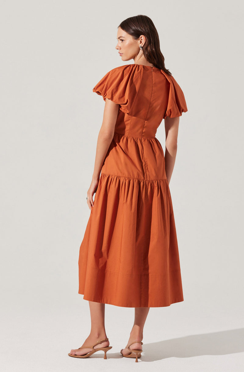 A woman is wearing a rust-colored dress with puffed short sleeves, a fitted waist, and a gathered, full-length skirt. The dress appears to be made of a lightweight fabric with a slight sheen. She is also wearing nude, strappy heeled sandals.