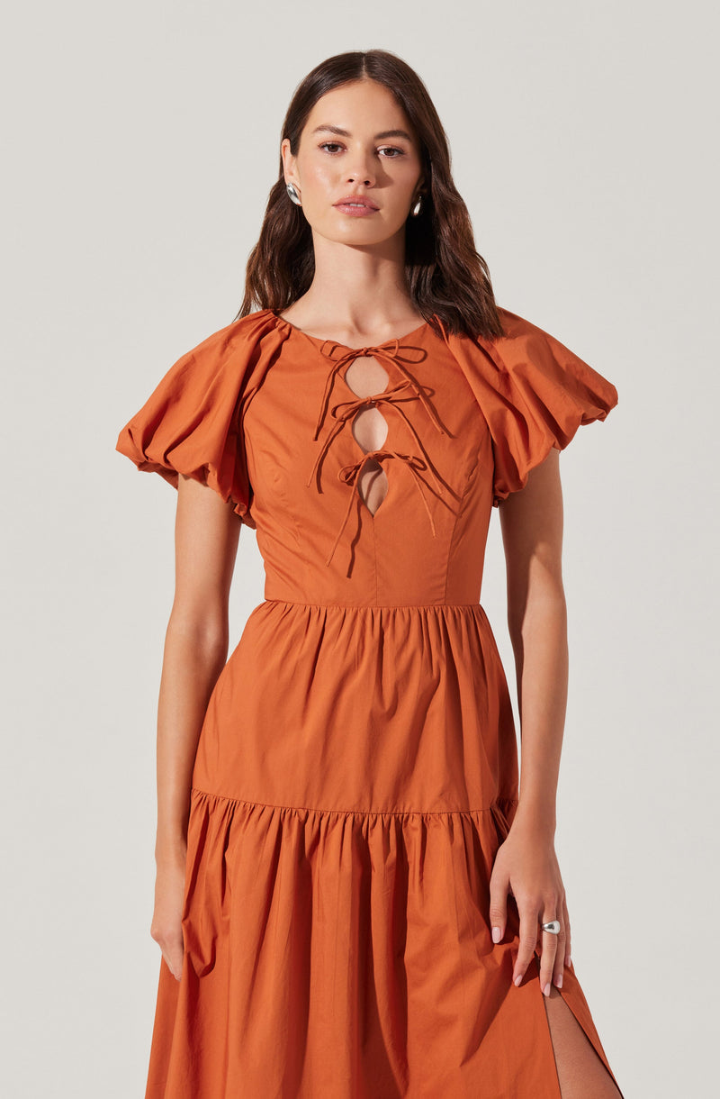 A woman is wearing a burnt orange dress with puffed short sleeves, a fitted waist, and a tiered skirt. The dress has a round neckline with a keyhole opening in the front, accented by a drawstring tie that creates a bow.