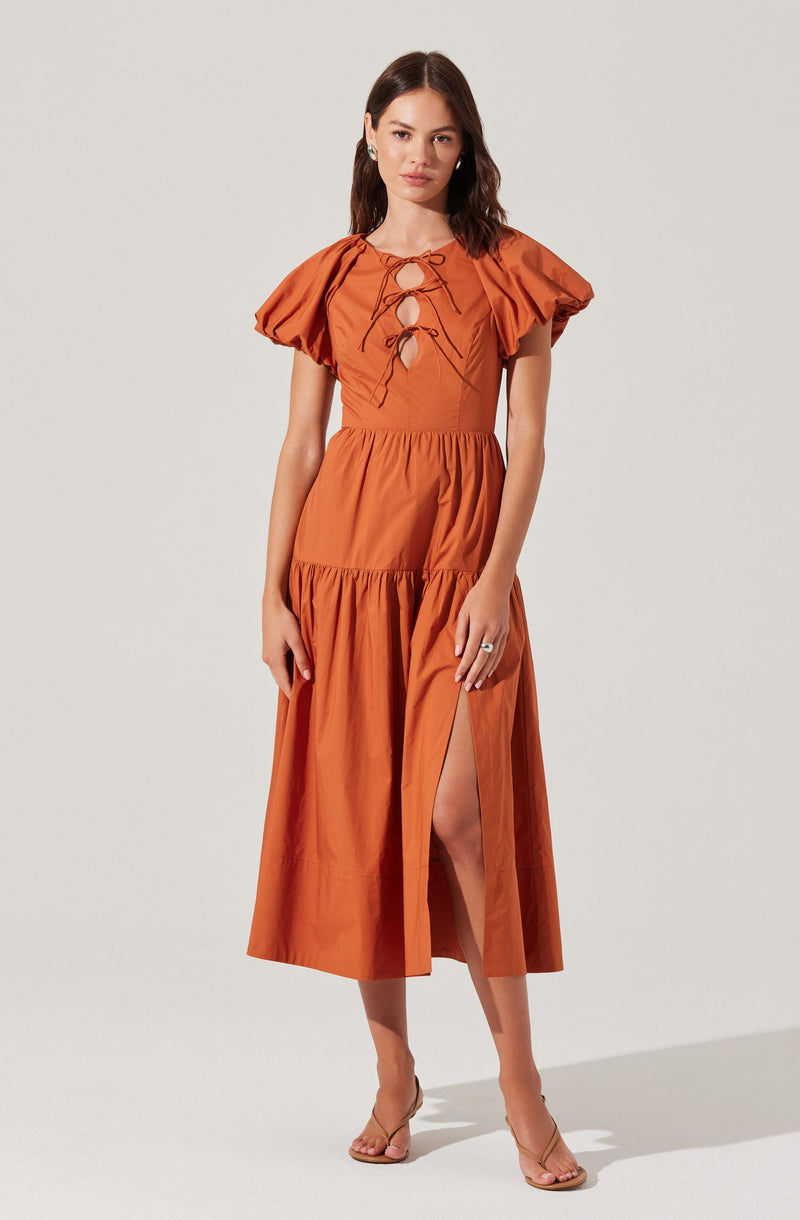 A woman is wearing an orange maxi dress with puffed short sleeves and a fitted waist. The dress features a lace-up detail with ties at the neckline and a high slit on one side, revealing her leg. She is also wearing nude high-heeled sandals and a ring on her right hand.