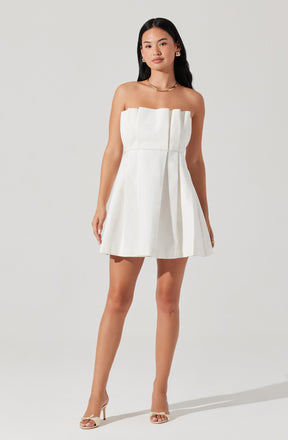 A woman is wearing a strapless white dress with a pleated skirt. The dress has a straight neckline with a ruffled detail at the top. She is accessorized with gold jewelry, including hoop earrings, a necklace, and rings. She is also wearing strappy high-heeled sandals.