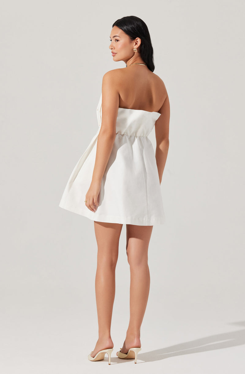 A woman is wearing a strapless white mini dress with a gathered waist. She has on beige high-heeled shoes with a pointed toe and stiletto heel. The dress appears to be made of a light, structured fabric.