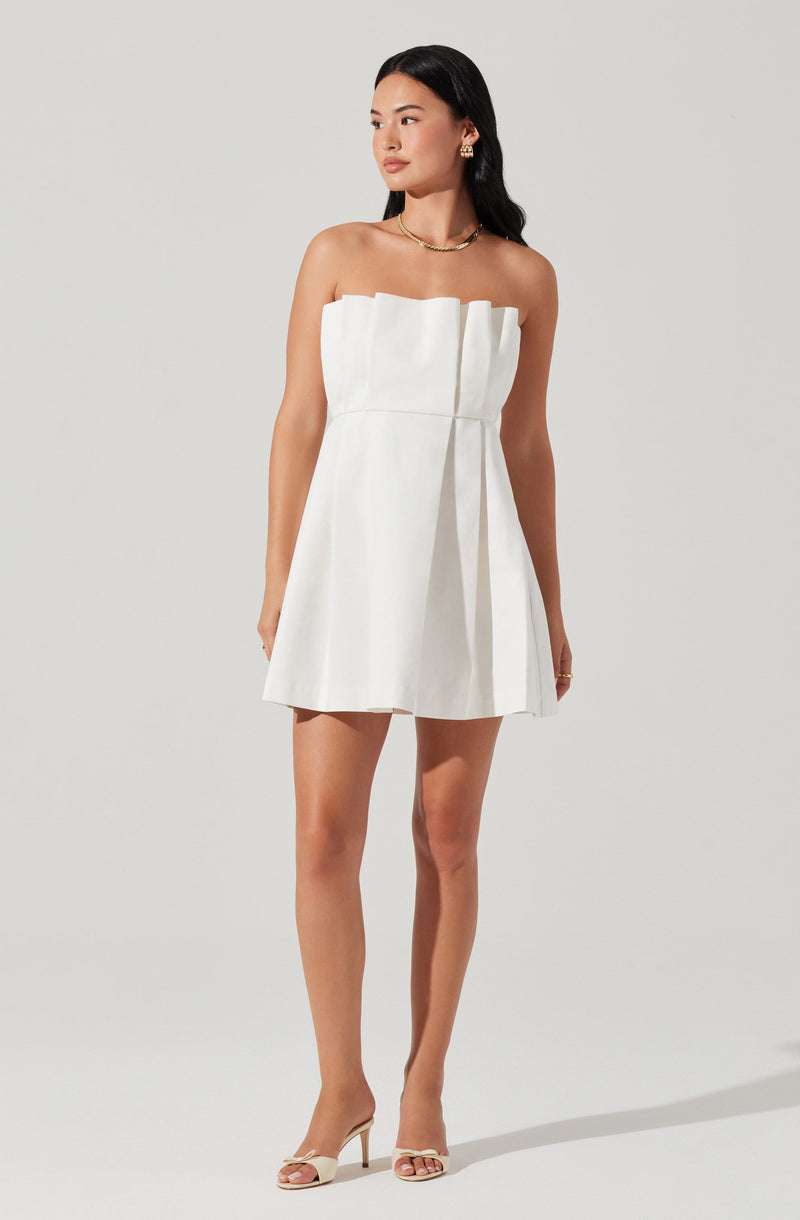 A woman is wearing a strapless white dress with a structured, pleated bodice and a slightly flared skirt. She has paired the dress with nude high-heeled sandals, a gold necklace, gold earrings, and a ring.