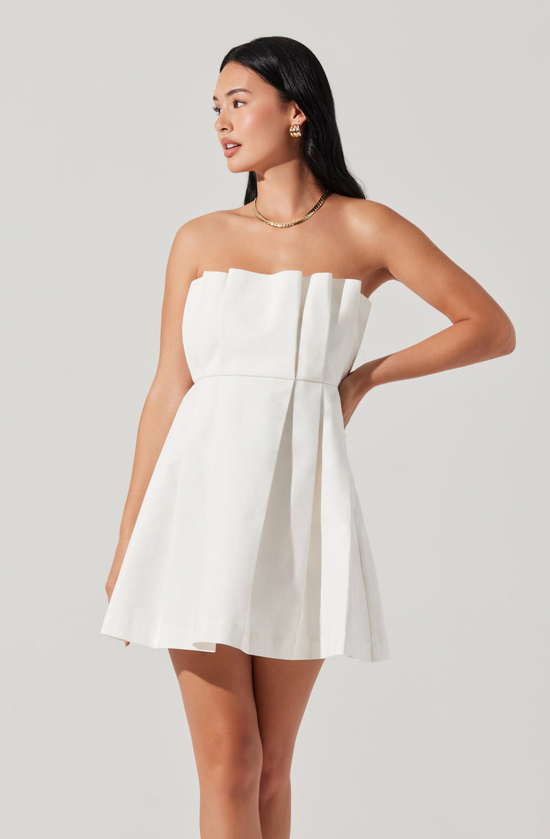 A woman is wearing a strapless, white dress with vertical pleats and a wide, pleated waistband that ties into a bow at the side. She accessorizes with gold earrings, a gold chain necklace, and has long, dark, straight hair.
