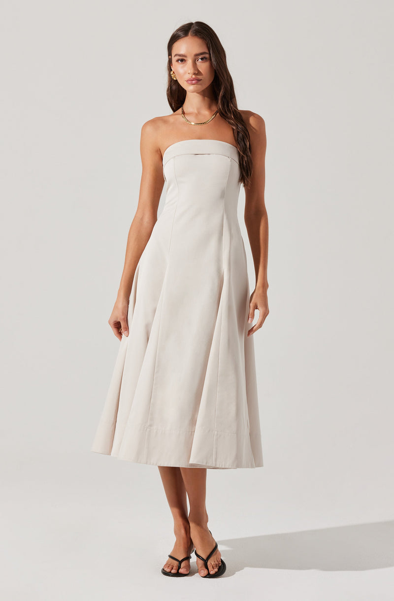 A woman is wearing a strapless, white, midi-length dress with a fitted bodice and a full, structured skirt. She has accessorized with gold earrings and a gold necklace, and is wearing black flip-flop style sandals.