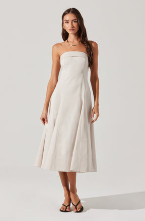 A woman is wearing a strapless, white, midi-length dress with a fitted bodice and a full, structured skirt. She has accessorized with gold earrings and a gold necklace, and is wearing black flip-flop style sandals.
