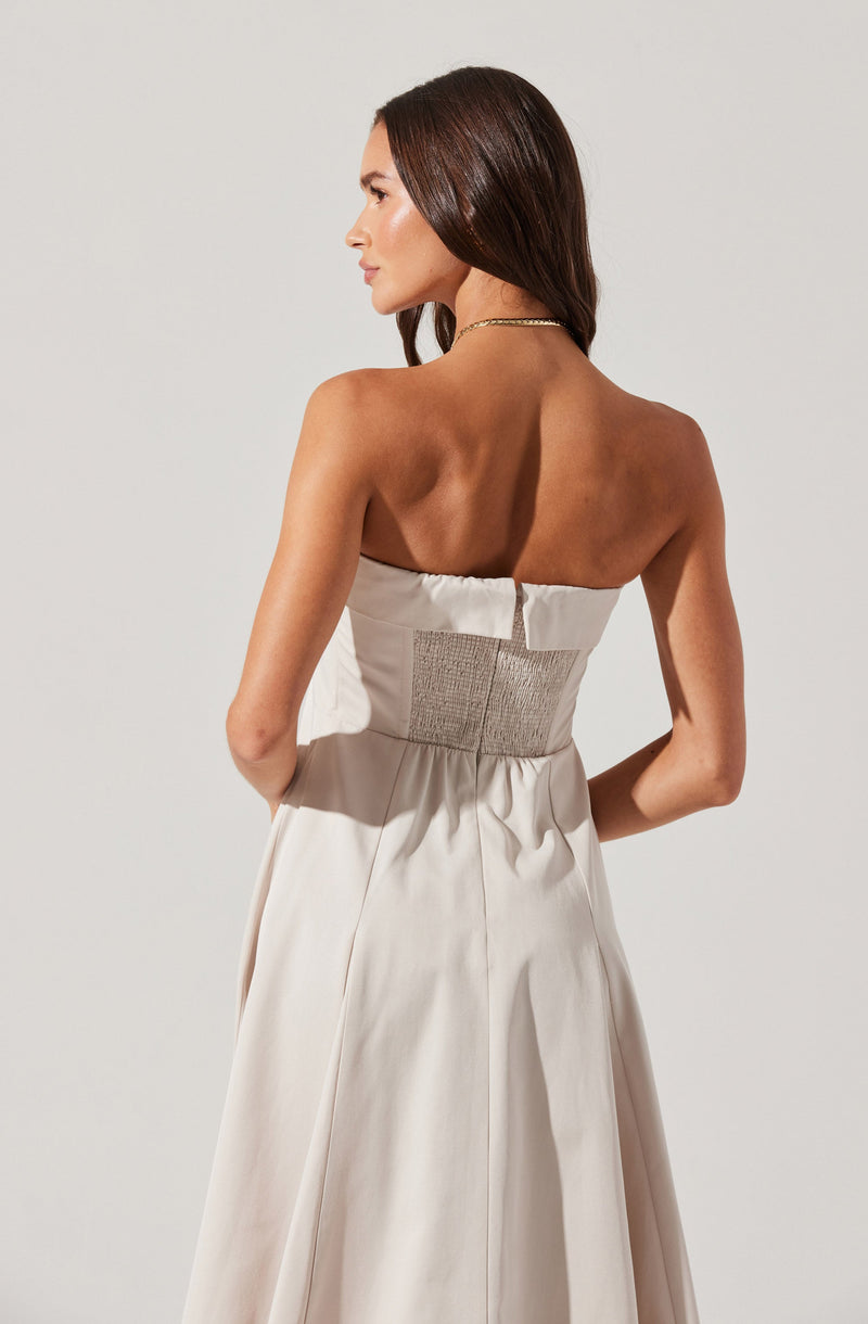 A woman wearing a strapless, off-white gown with a fitted, ruched waistband. The dress has a smooth, flowing skirt with pleats, and she is accessorized with a delicate necklace. Her hair is styled in loose waves.