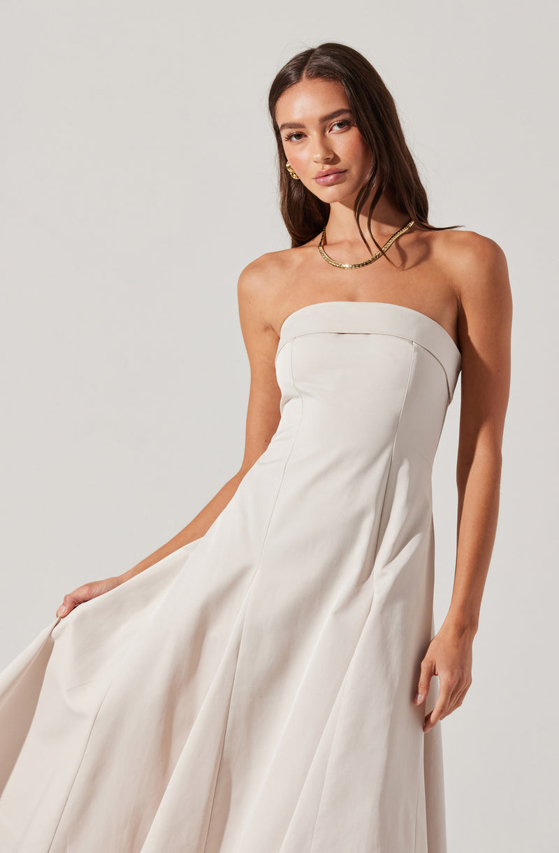 A woman is wearing a strapless white dress with a fitted bodice and a full, flowing skirt. She accessorizes with a gold necklace and matching gold earrings.