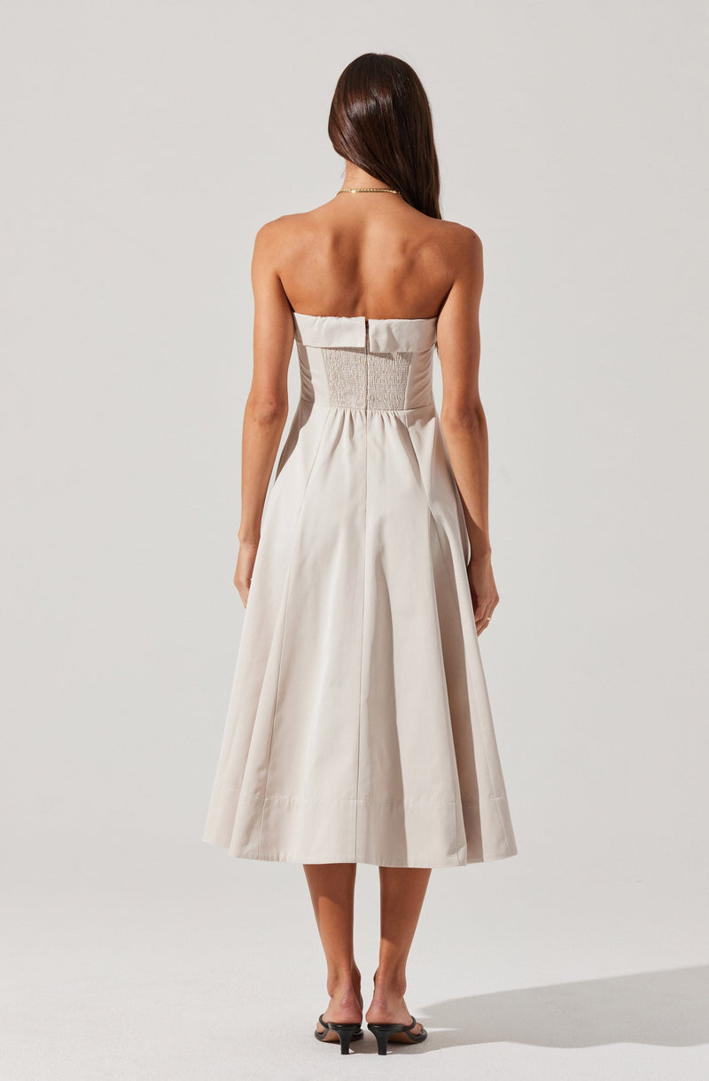A woman is wearing a strapless, knee-length dress in a light, neutral color. The dress features a fitted bodice with ruching and gathers at the waist, creating a draped effect. The skirt is full with pleats that give it volume and a subtle sheen. The woman is also wearing open-toe heeled sandals and a delicate necklace.